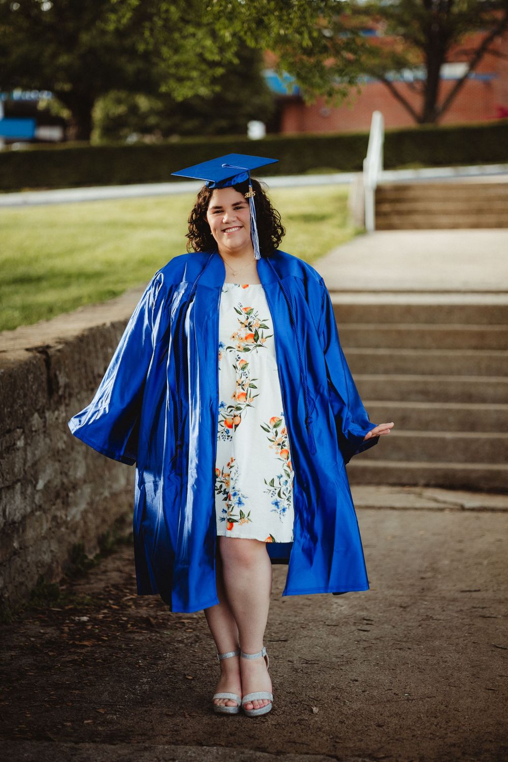 A graduate wearing a bright blue commencement gown and cap poses on outdoor steps while wearing a floral print dress.