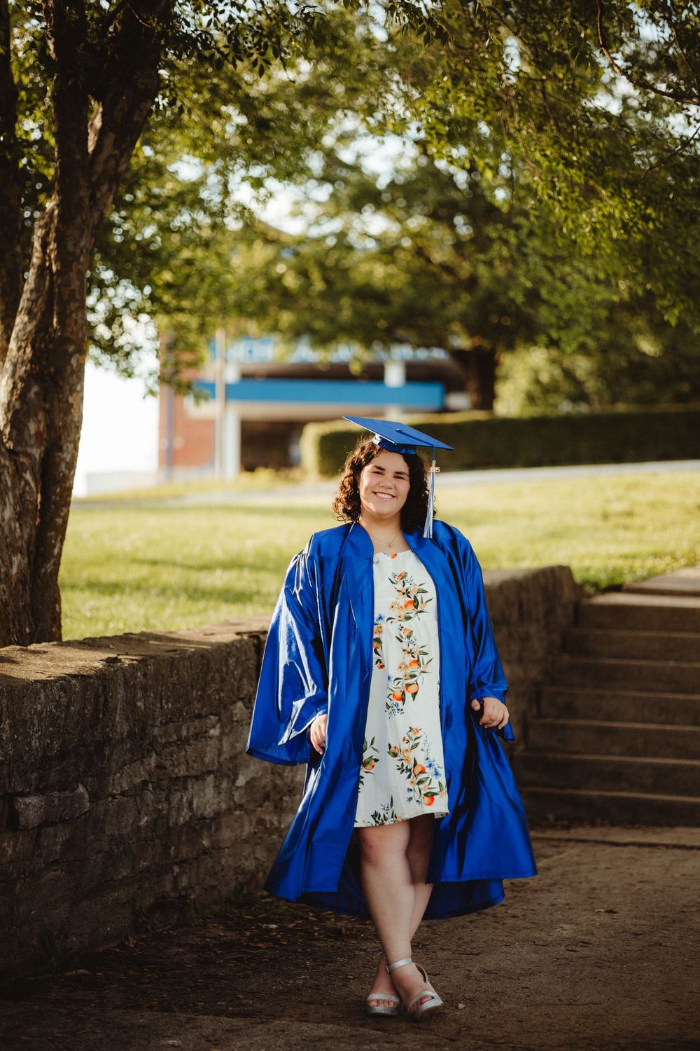 Graduate wearing bright blue cap and gown walks down brick steps on a sunny day under trees.