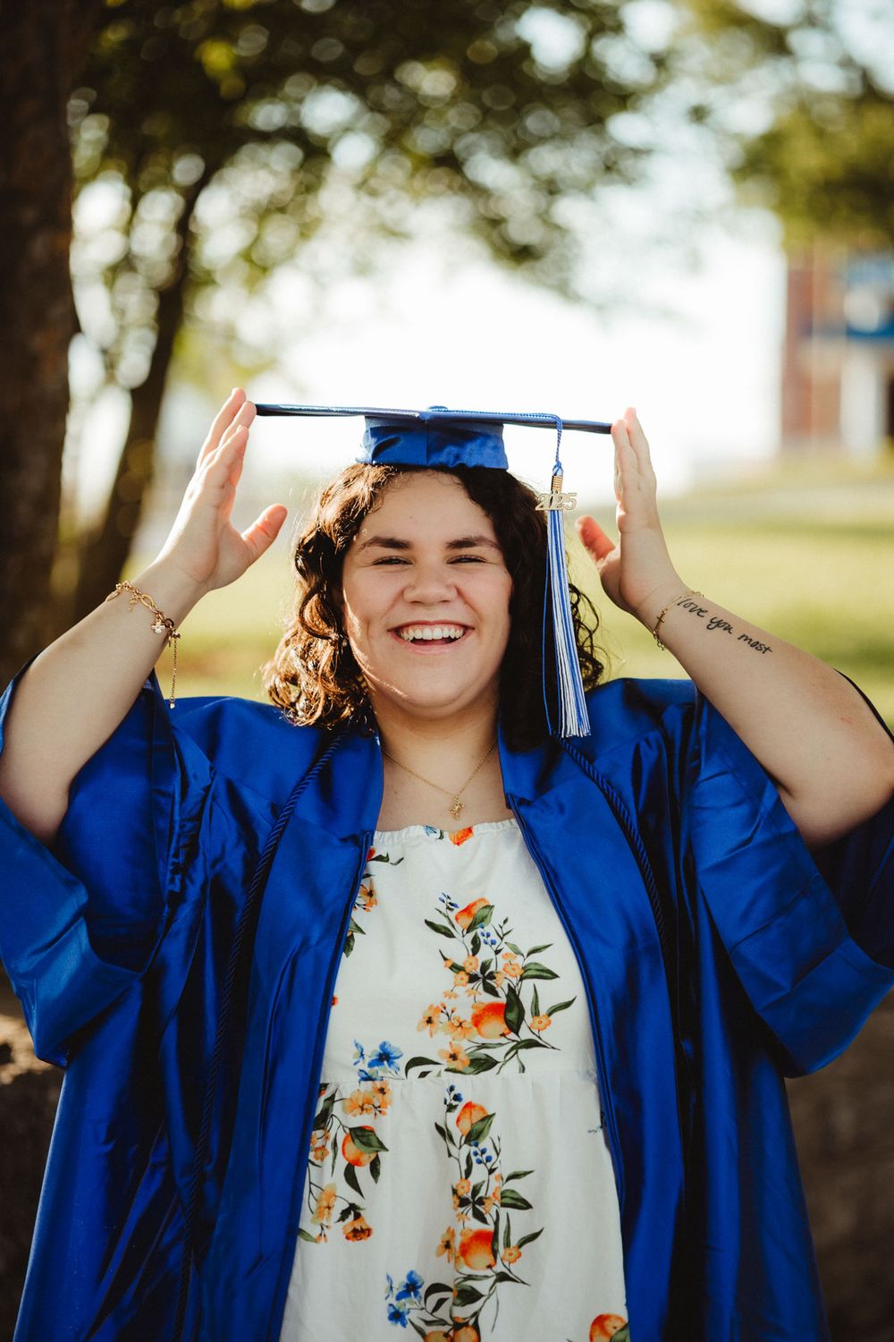 Graduate in bright blue cap and gown smiles joyfully while adjusting mortarboard during outdoor graduation photoshoot.