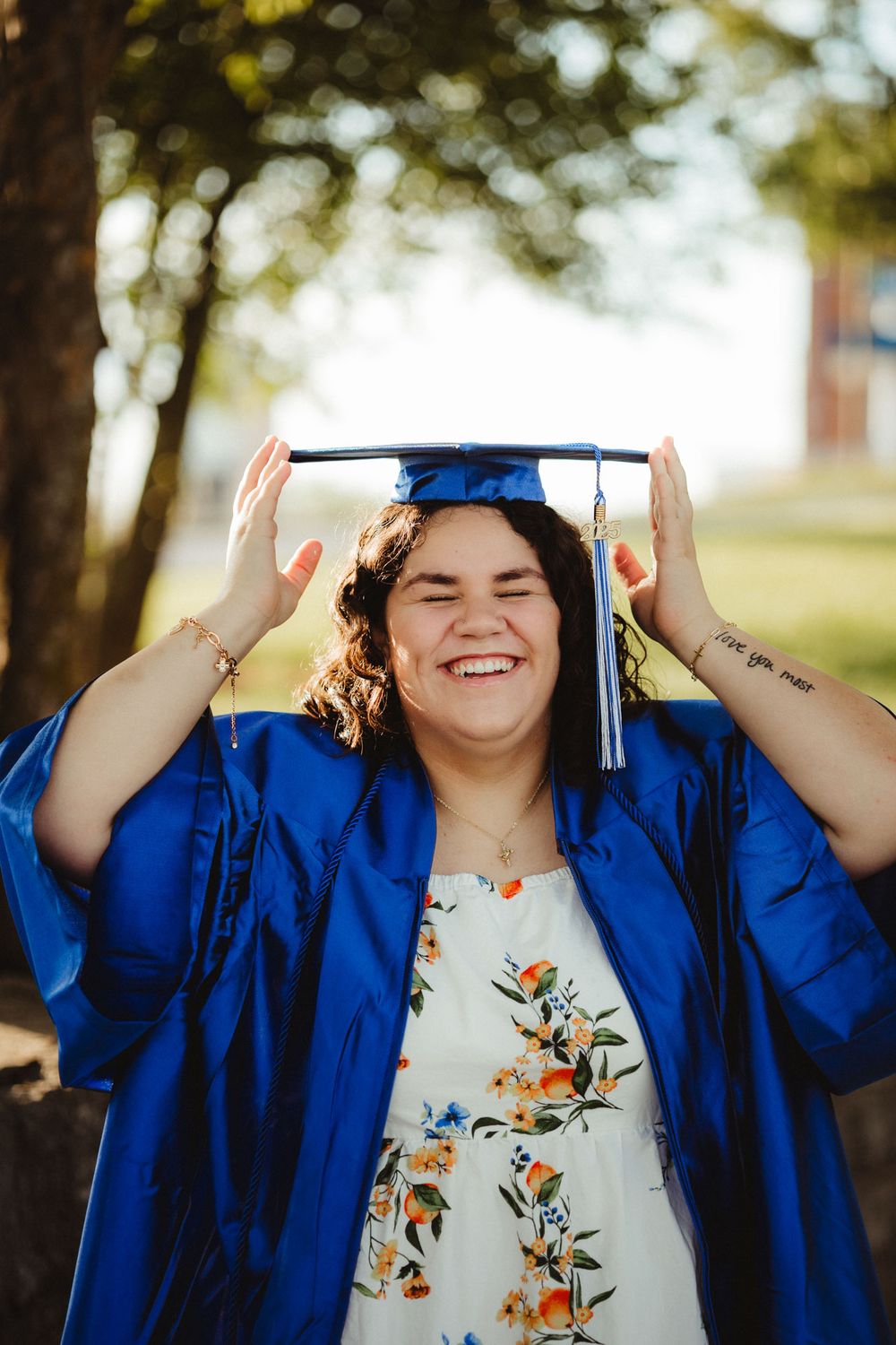 A graduate in bright blue cap and gown smiles joyfully while touching their mortarboard during an outdoor photo session.