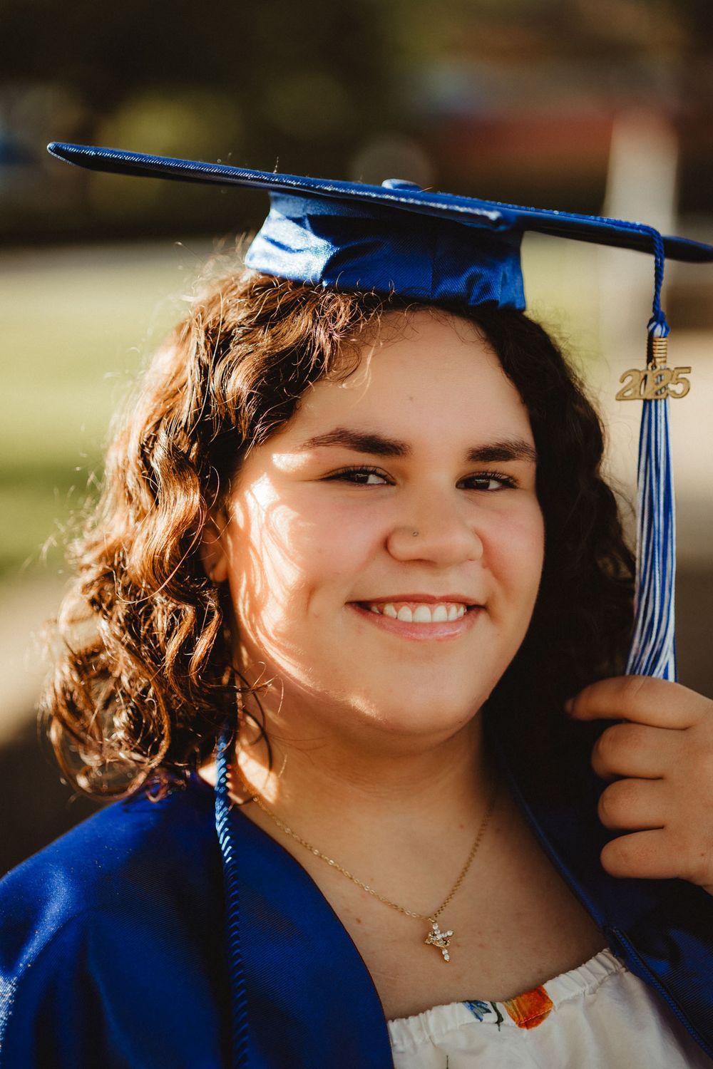 Graduate wearing a blue cap and gown with tassel and gold cross necklace smiles during commencement ceremony.