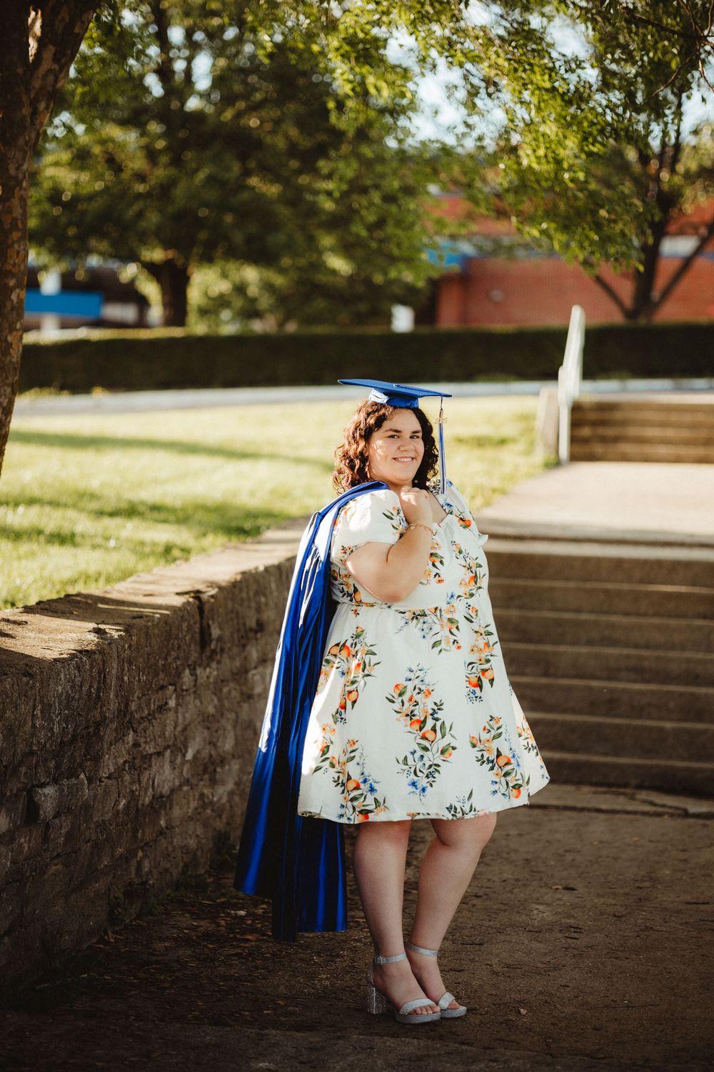 Graduate in a blue cap and gown with floral dress poses on stone steps during sunny outdoor photo session.