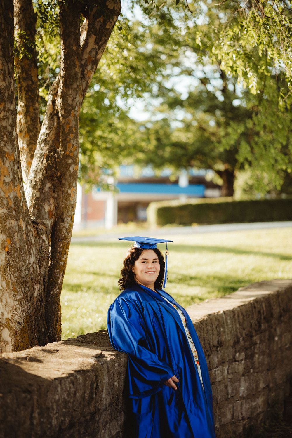 Graduate in traditional blue cap and gown stands by stone wall under tree in sunlit outdoor setting.