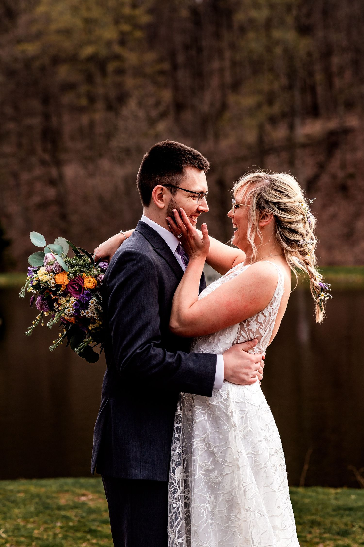 A bride and groom share an intimate moment by a lake with the bride holding a colorful wildflower bouquet.