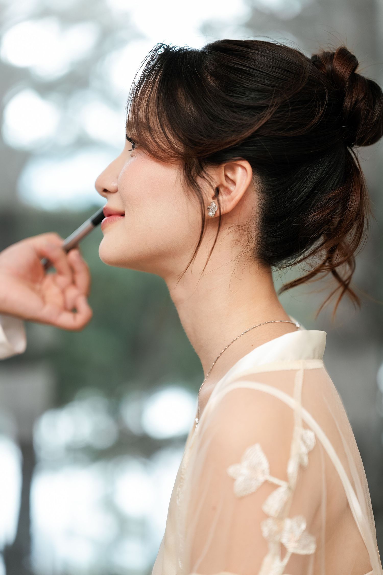 Side profile of someone having makeup applied while wearing a delicate floral embroidered sheer white dress.