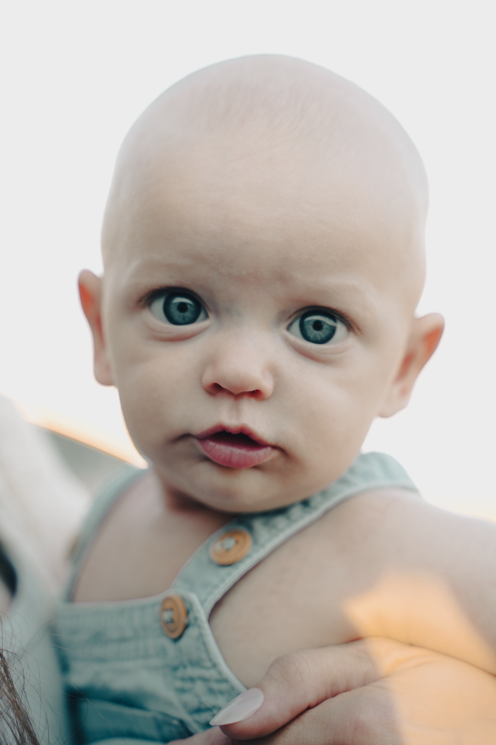 Close-up portrait of baby in denim overalls with striking blue eyes against light background.