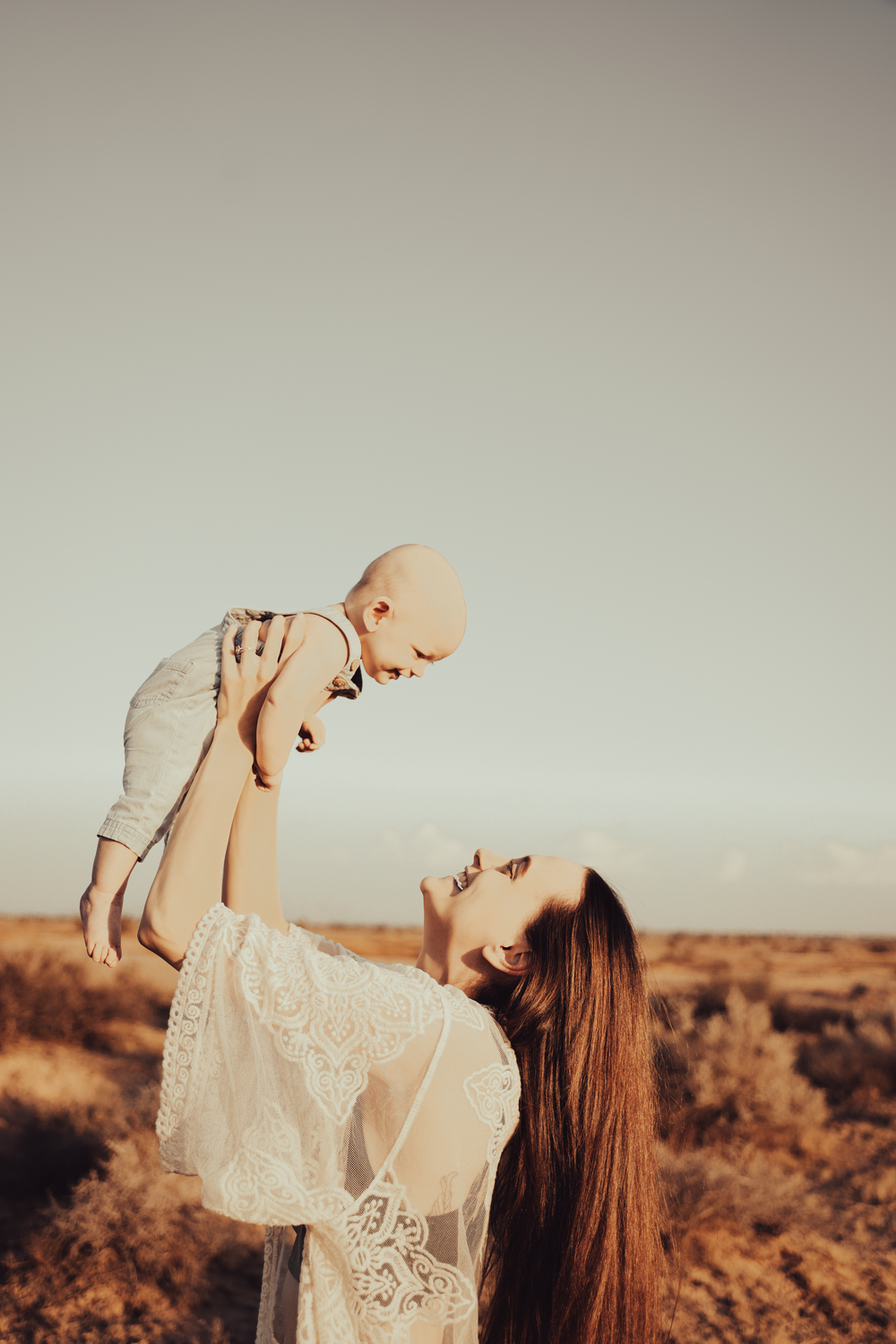 A mother in a white lace dress lifts her baby against a golden sunset in an outdoor setting.
