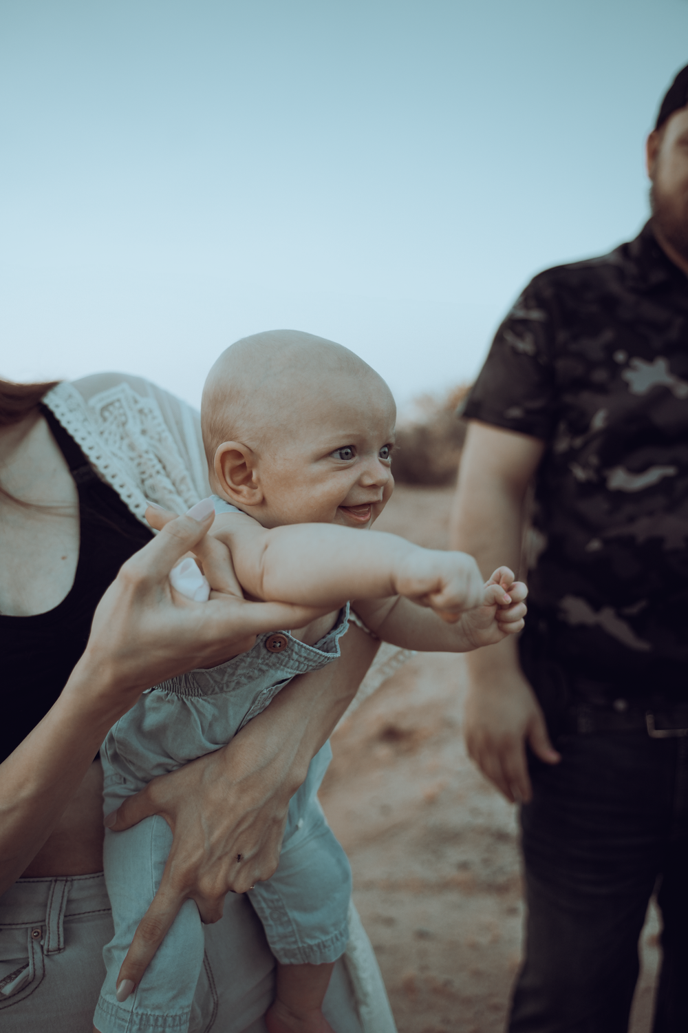 A happy baby in gray overalls smiles while being held outdoors.