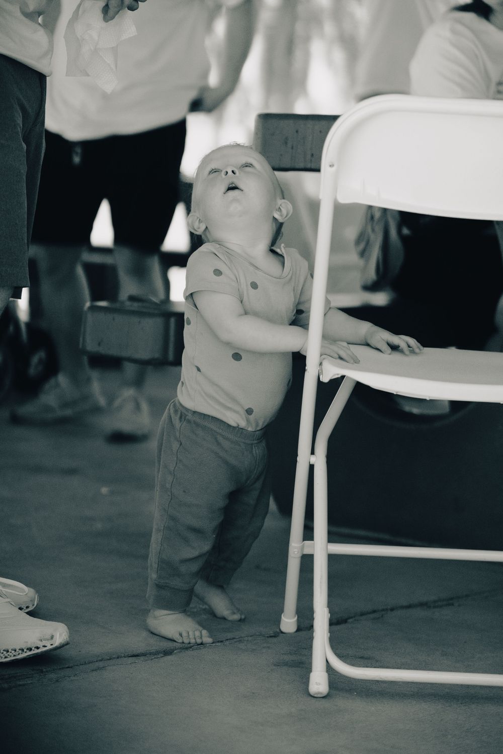 A small child leans against a white folding chair in black and white photography.