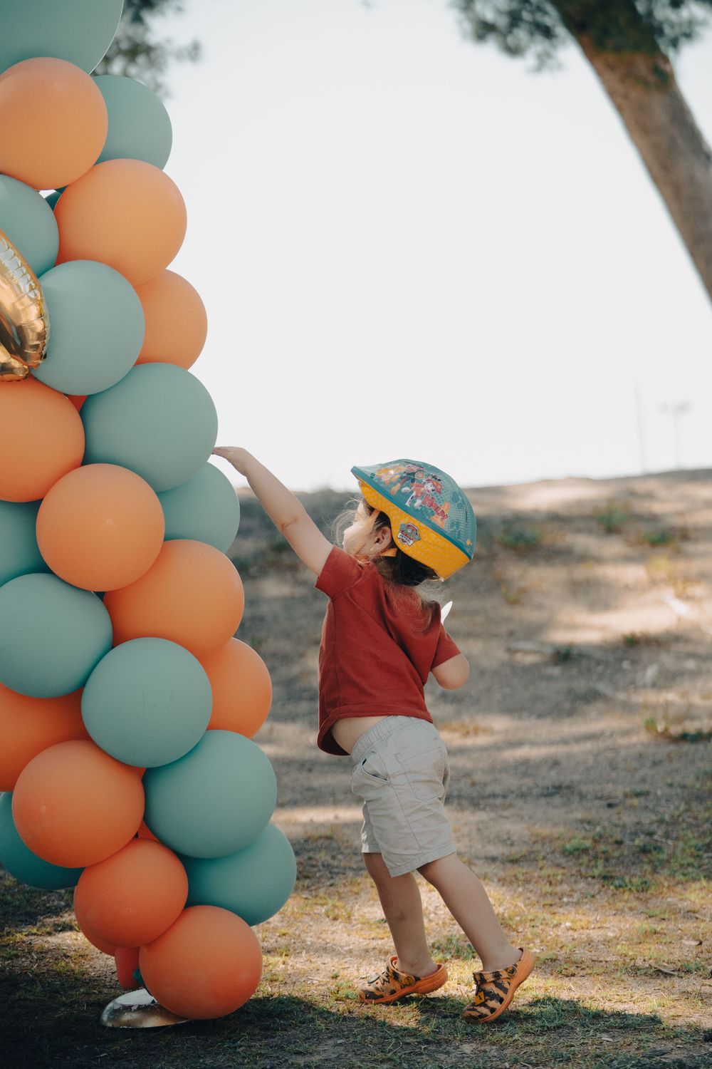 Small child reaching up to touch orange and blue balloon arch outdoors.