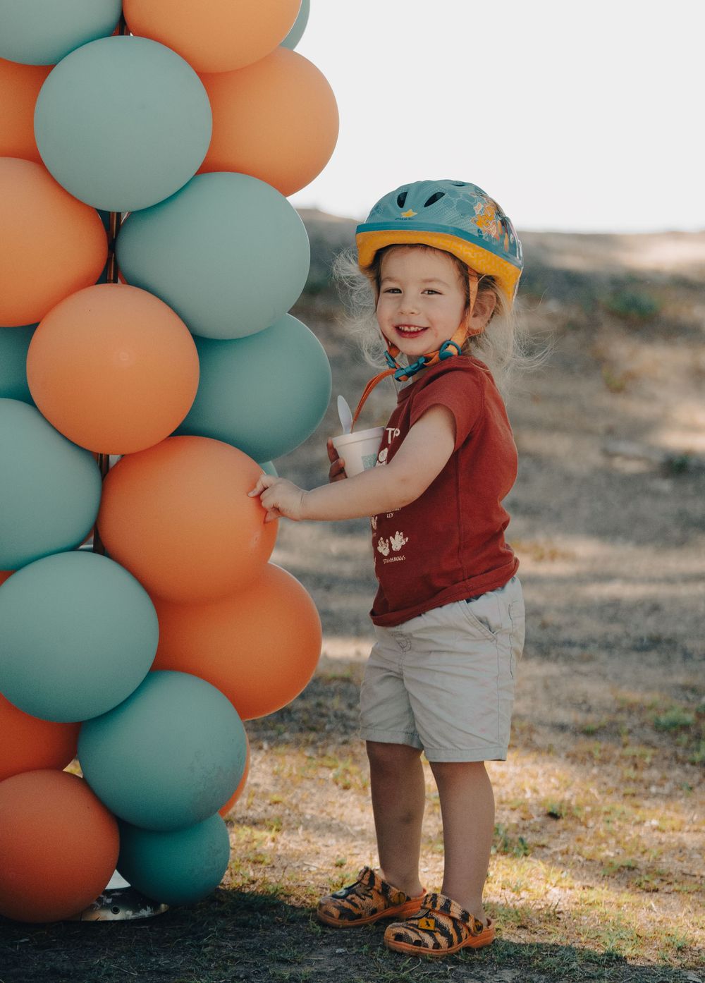 Child wearing red shirt and yellow helmet plays with colorful balloon display.