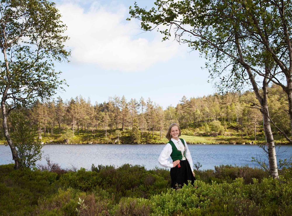 Jente stående foran et vann i bunad og smiler til fotografen under konfirmantfoto i Lyngdal