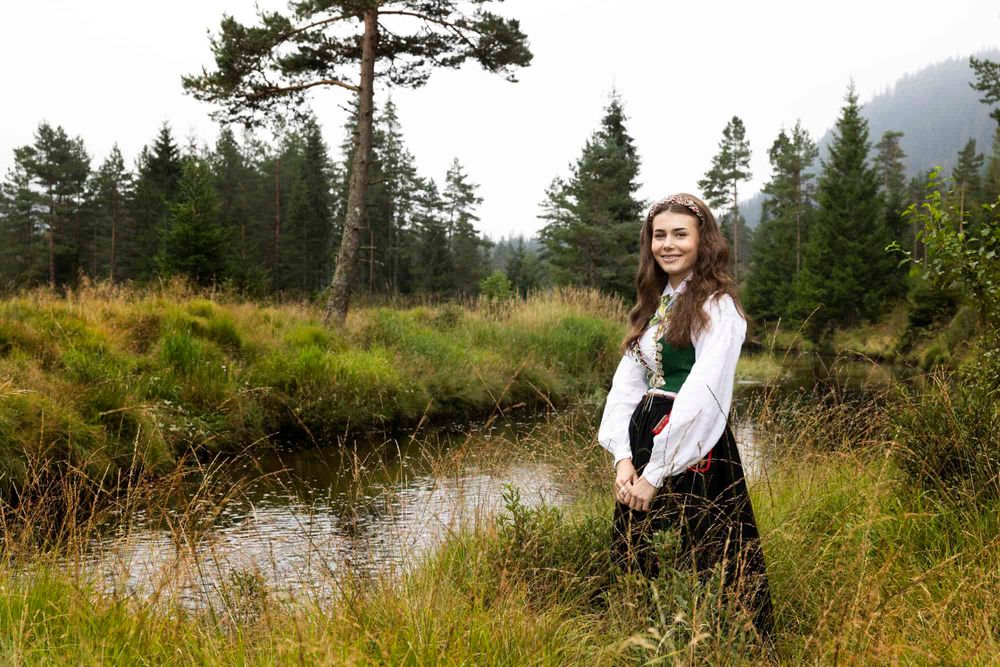 Jente stående foran et vann i bunad og smiler til fotografen under konfirmantfoto i Lyngdal