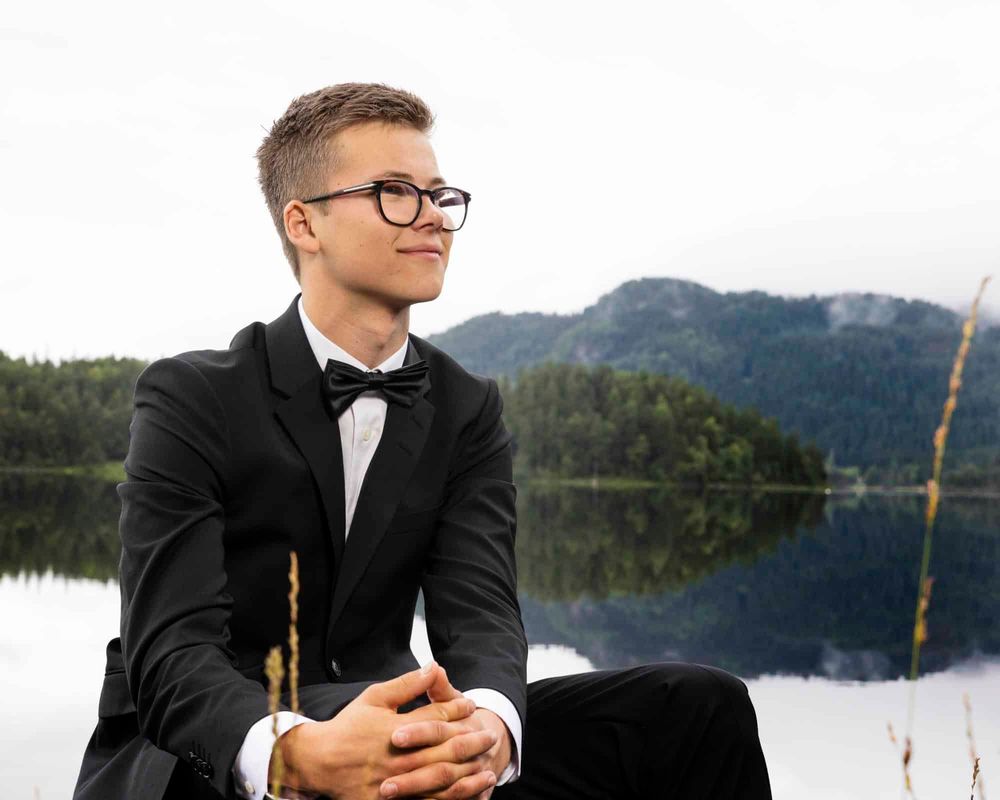 Konfirmant i dress med briller sittende foran en vann i Lyngdal