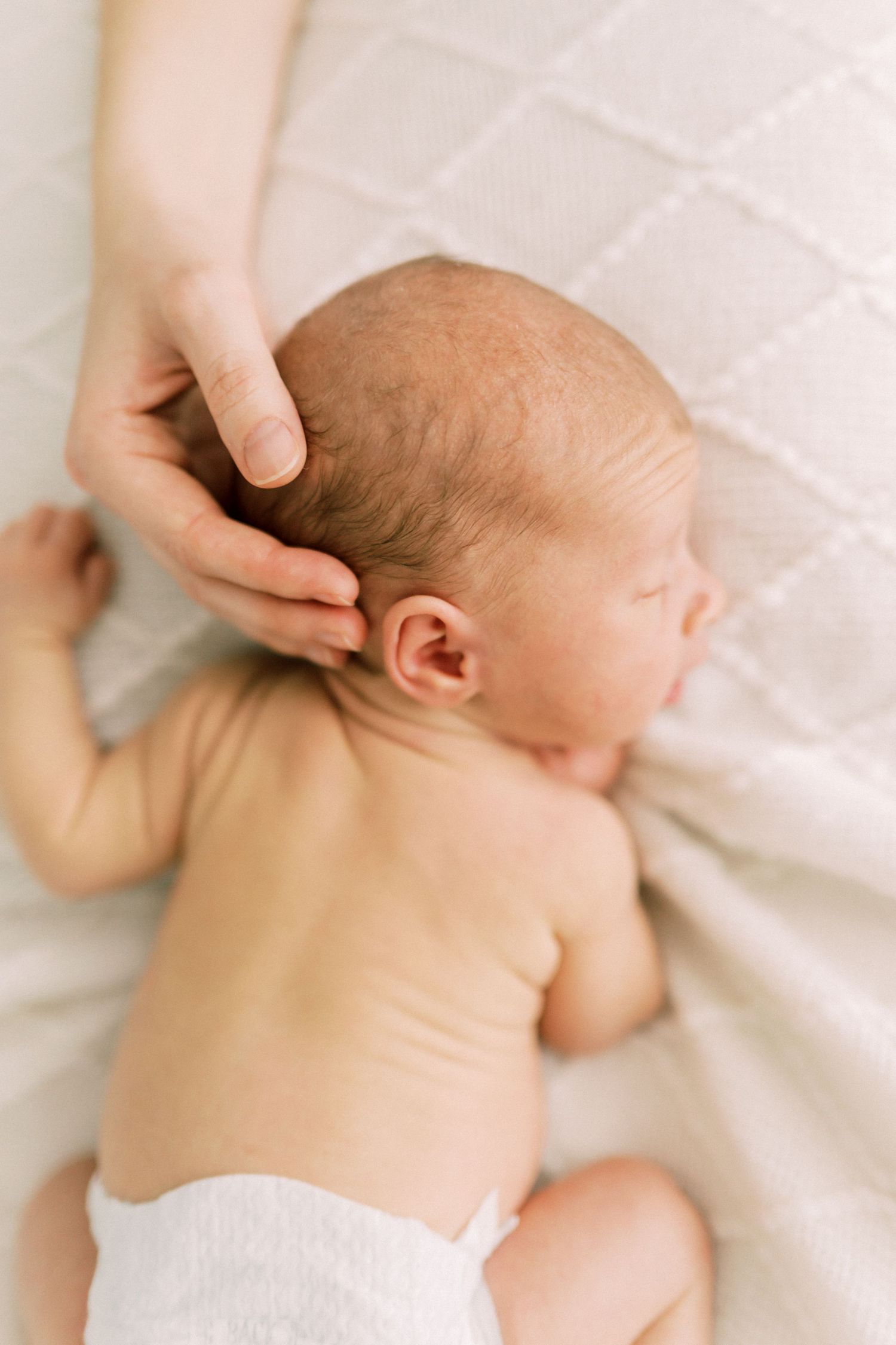 A gentle hand caresses a newborn baby in white diaper lying on soft white bedding.