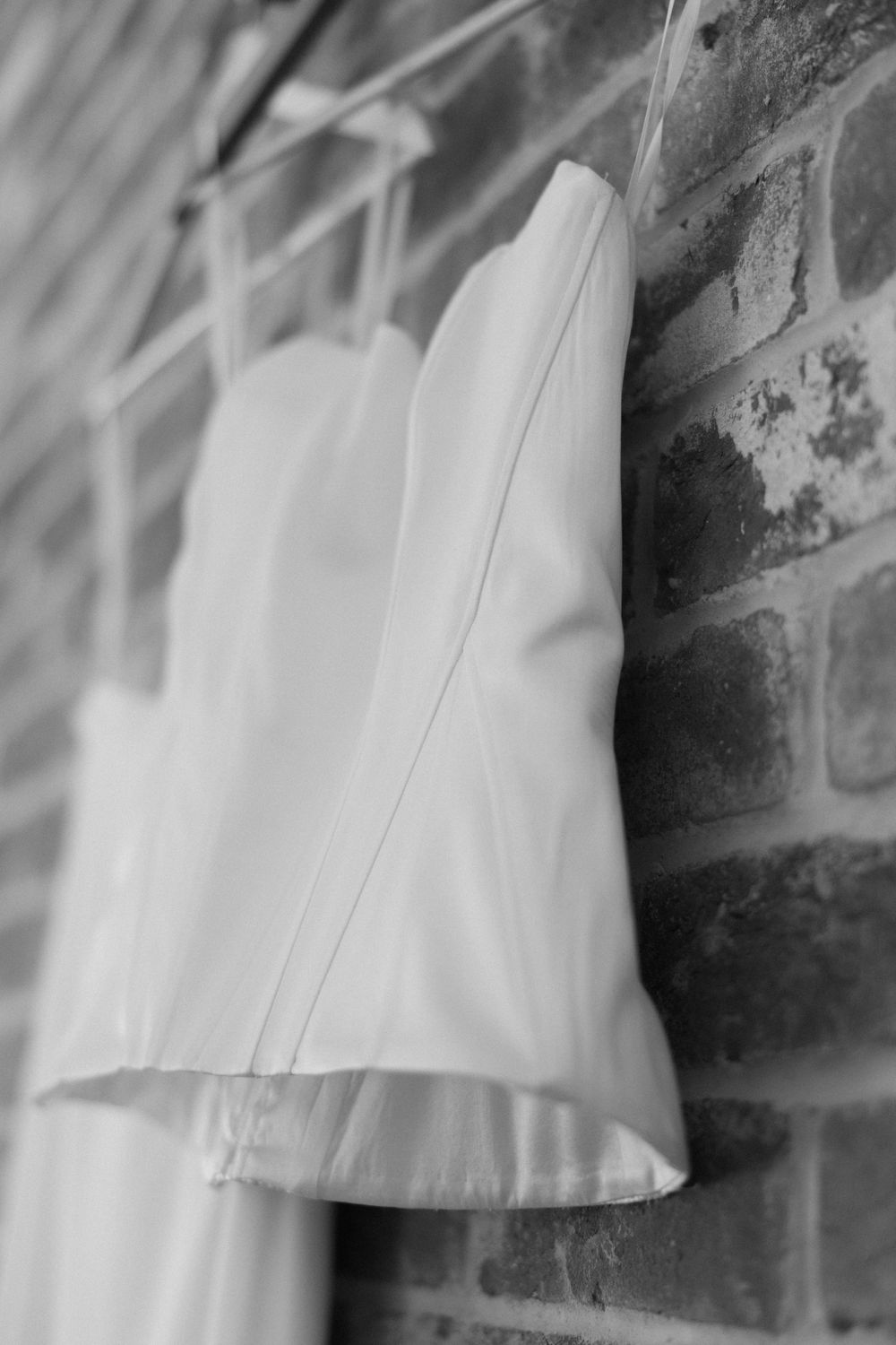 A white wedding dress hanging against an exposed brick wall in black and white photography.
