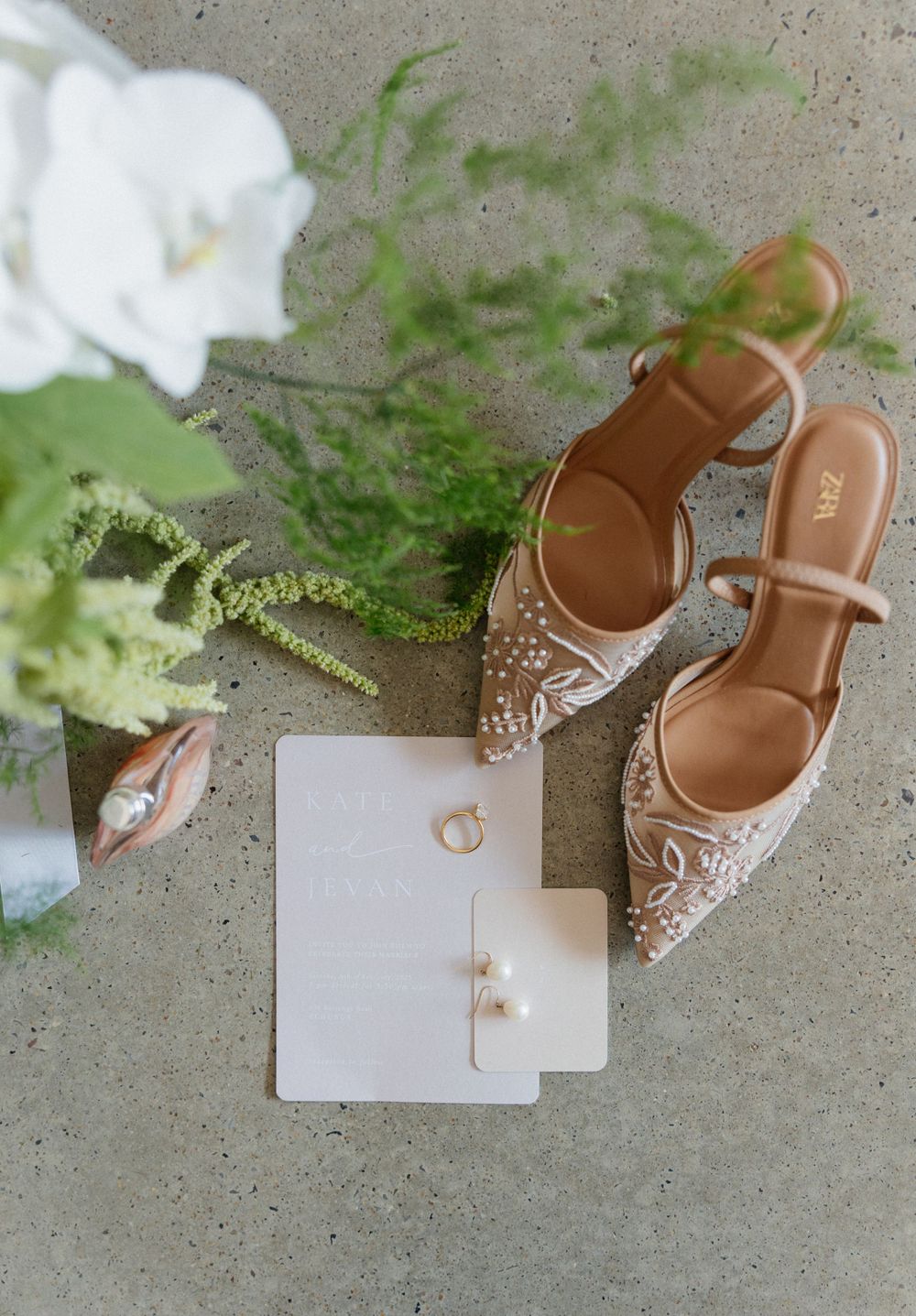 Elegant bridal details including sparkly pointed-toe heels, white florals, and wedding stationery arranged on concrete.