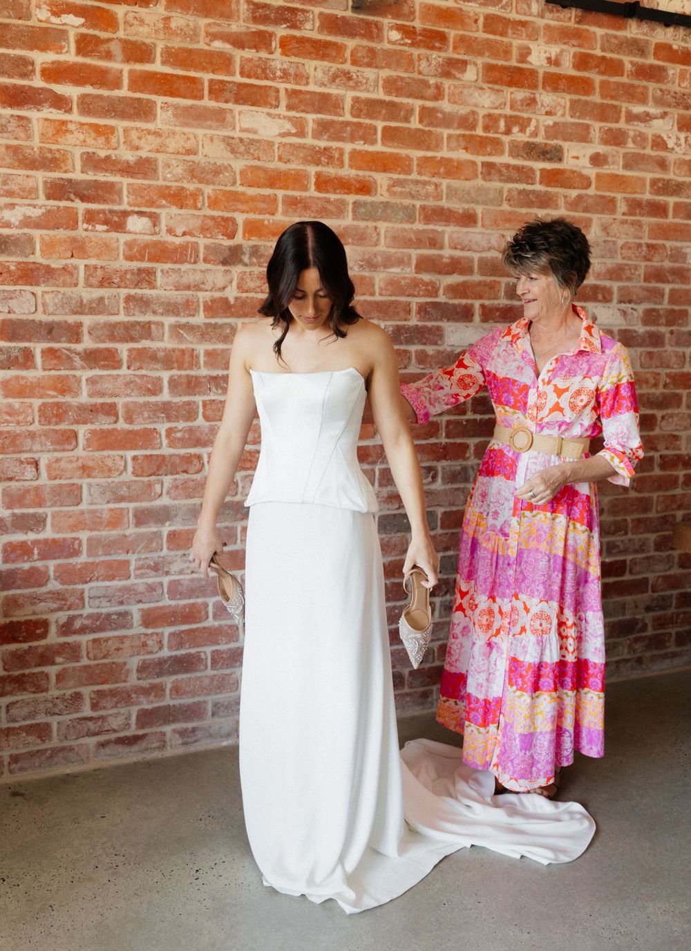 Two people stand by a brick wall, one wearing a white wedding dress while the other assists in a pink floral dress.