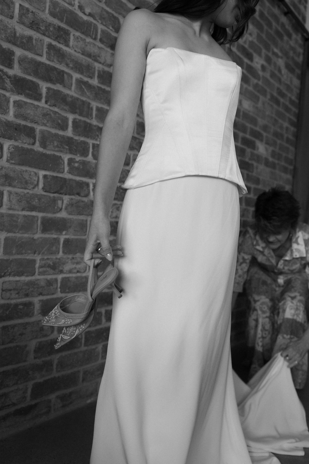 A black and white photograph shows an elegant white two-piece wedding dress with a fitted bodice against a brick wall.