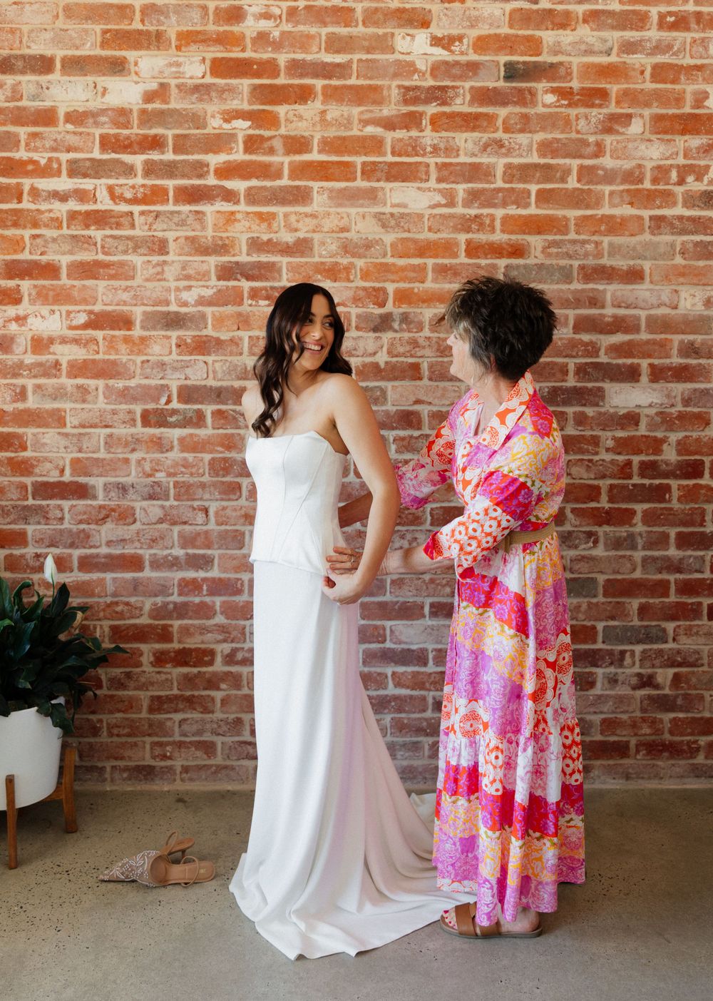 Two people stand in front of a brick wall, one wearing a white wedding gown and another in a vibrant pink floral dress.