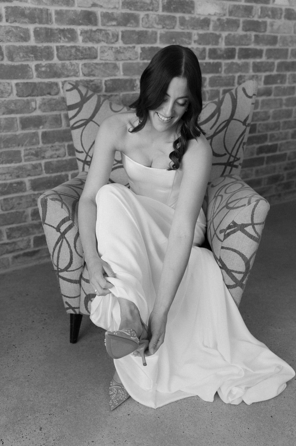 Black and white photo of a bride in a strapless gown adjusting her dress while sitting in a patterned armchair.