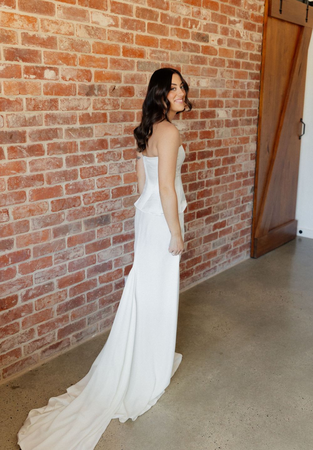A sleek white wedding gown with a fitted silhouette and train is modeled against a rustic brick wall backdrop.
