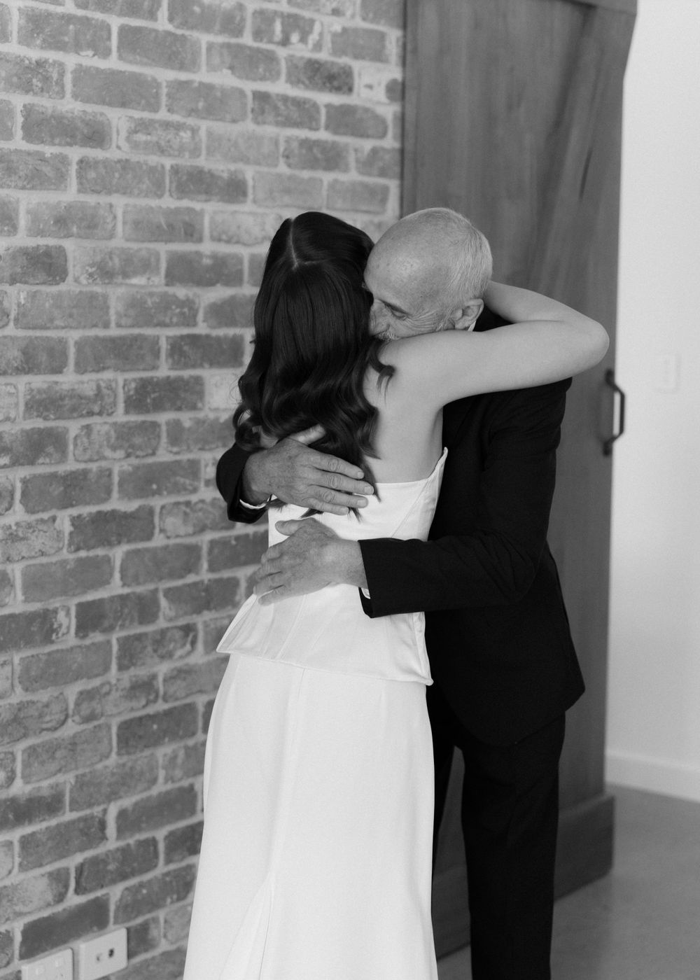 A tender moment between a couple embracing in formal wedding attire against a rustic brick wall.