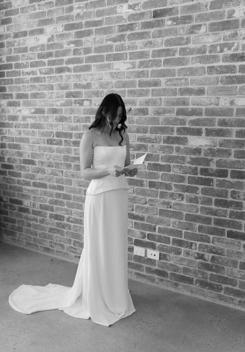 A black and white photograph of someone in an elegant white wedding gown reading against a rustic brick wall.