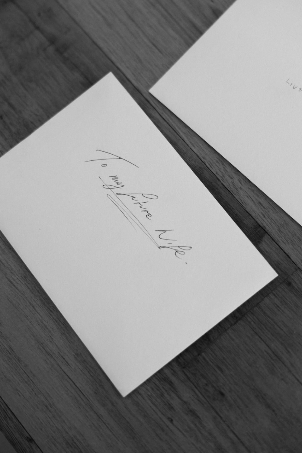 A black and white close-up shot of handwriting on a white piece of paper against a wooden surface.