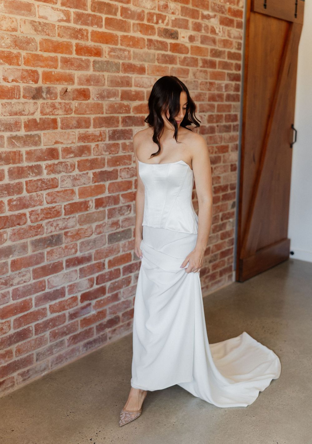 Elegant strapless white wedding gown with fitted silhouette and trailing train against rustic brick wall backdrop.