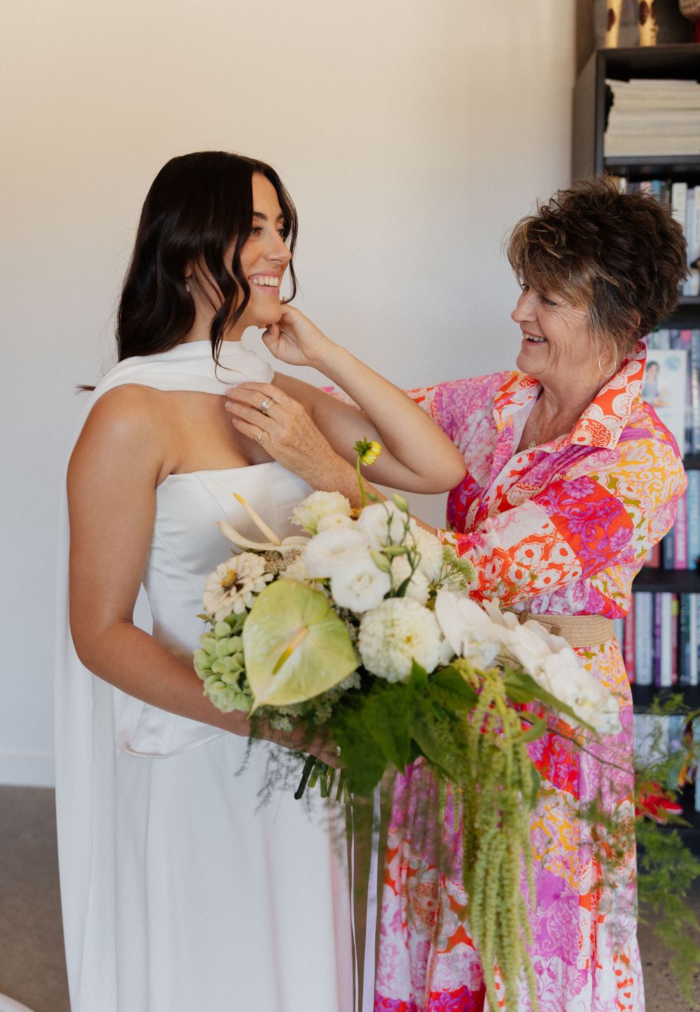 Two people share a tender moment at a wedding, one wearing white and holding a bouquet while another wears a floral dress.