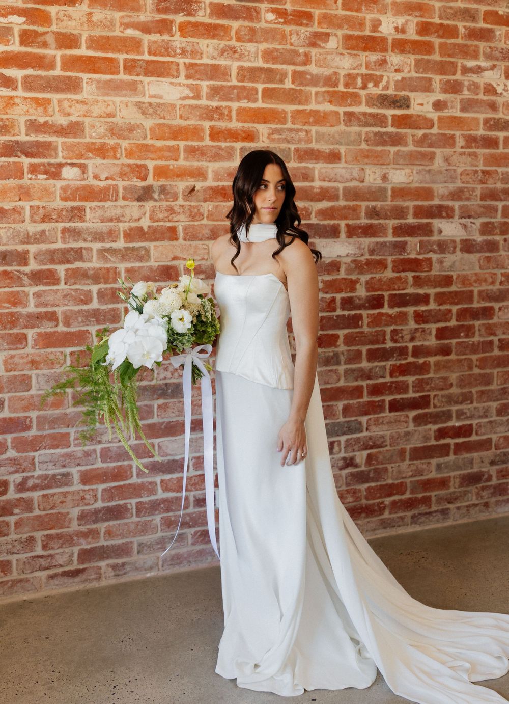 An elegant white wedding gown with a flowing train stands against a rustic brick wall as a floral bouquet adds natural beauty.