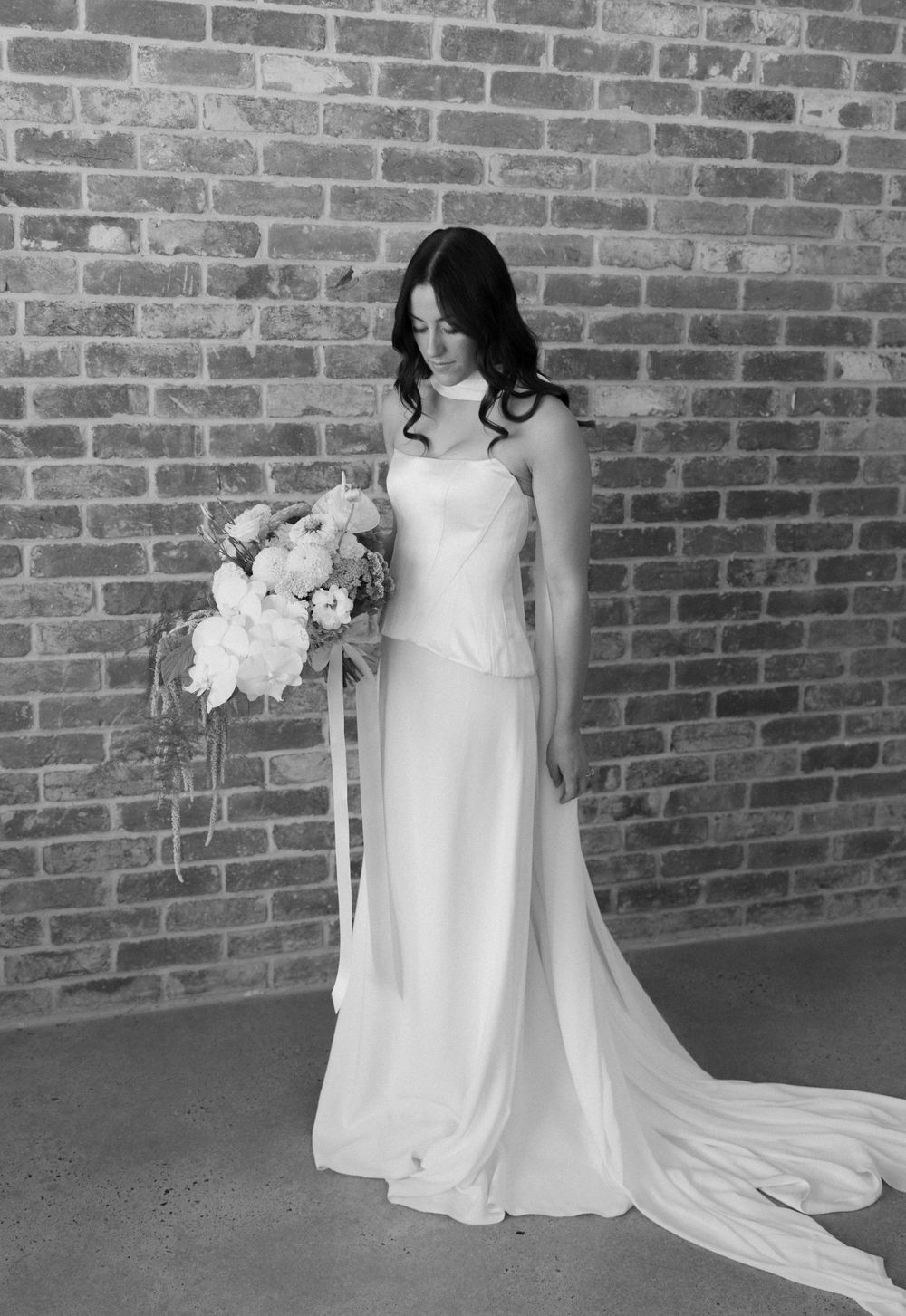 Black and white photo of bride in elegant strapless wedding dress holding bouquet against exposed brick wall.