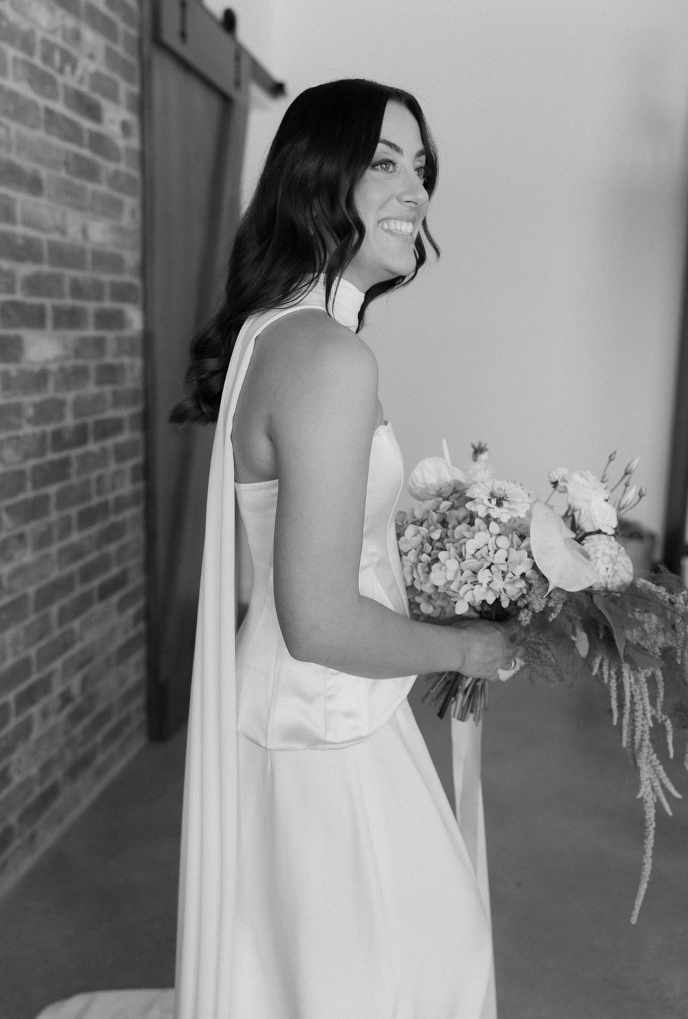 A person in an elegant white wedding dress holds a lush bouquet while standing by a brick wall.