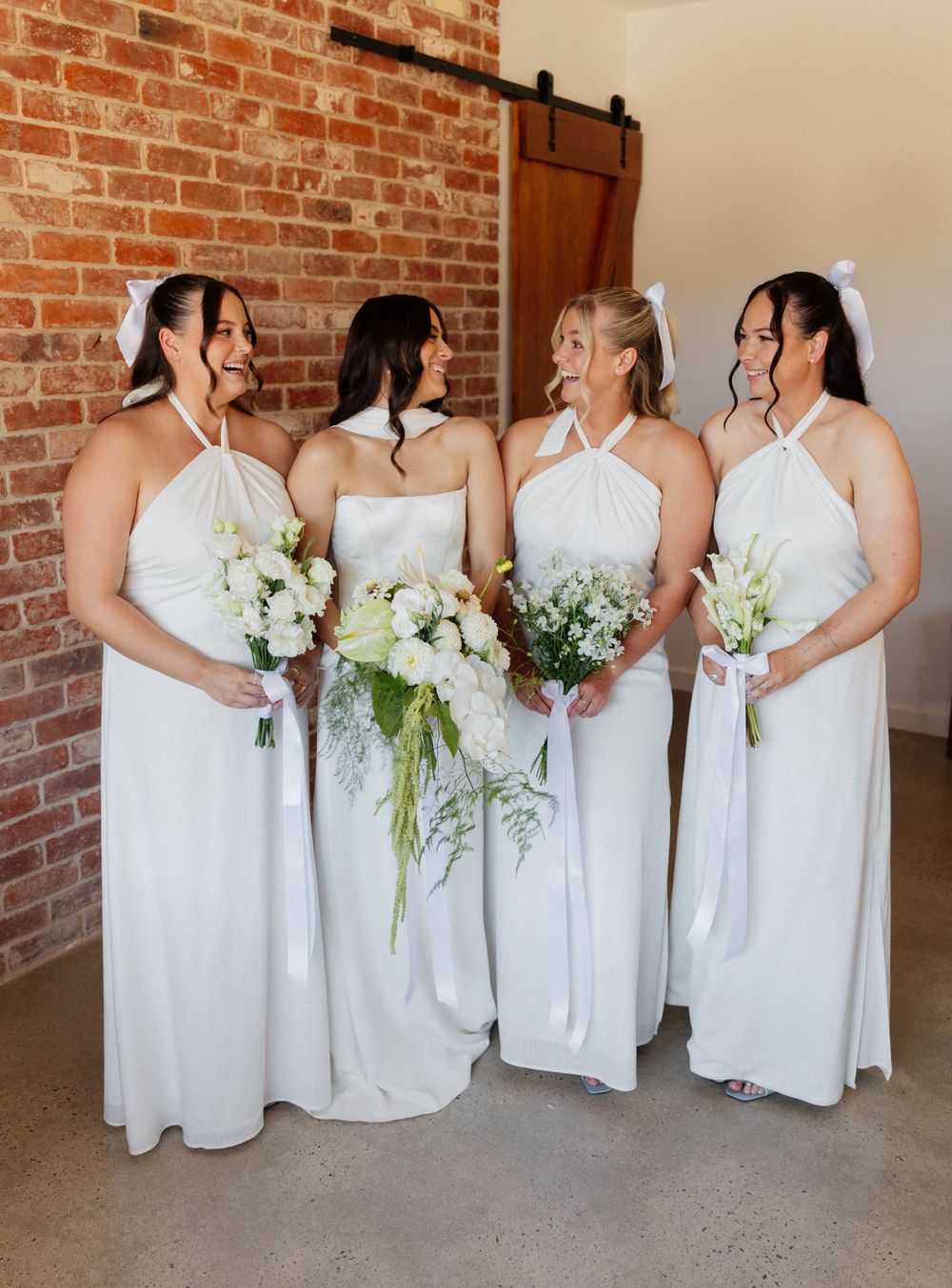 Bridesmaids in matching white halter gowns hold elegant white floral bouquets against exposed brick wall.