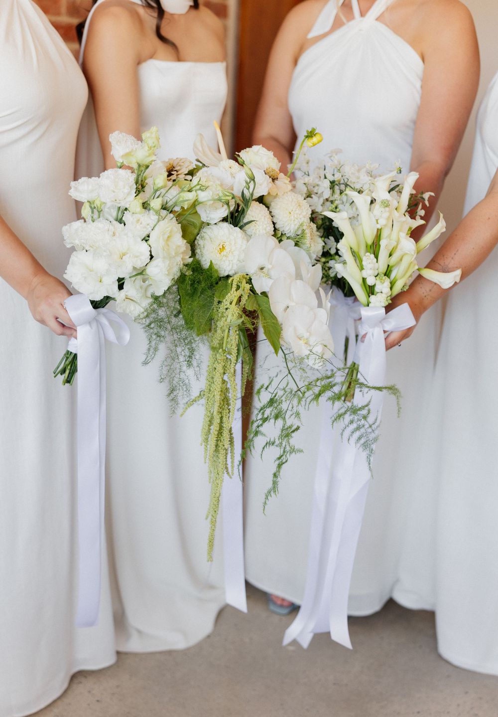 Elegant white wedding bouquets featuring fresh peonies, roses and delicate ferns held against flowing white bridesmaid dresses.