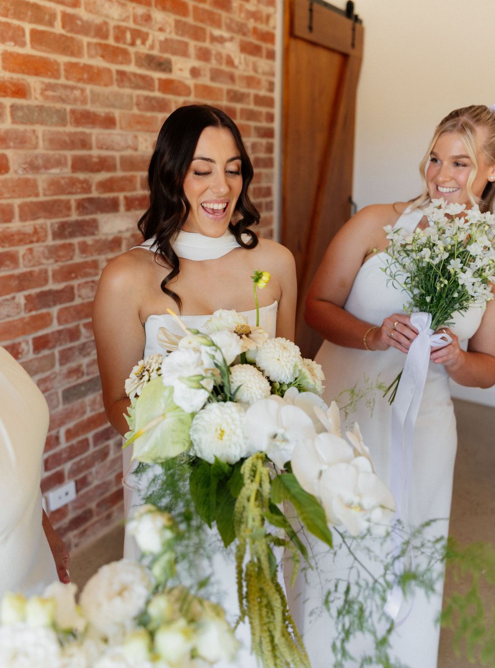 Bridal party members in white dresses hold beautiful white floral bouquets while standing against exposed brick wall.