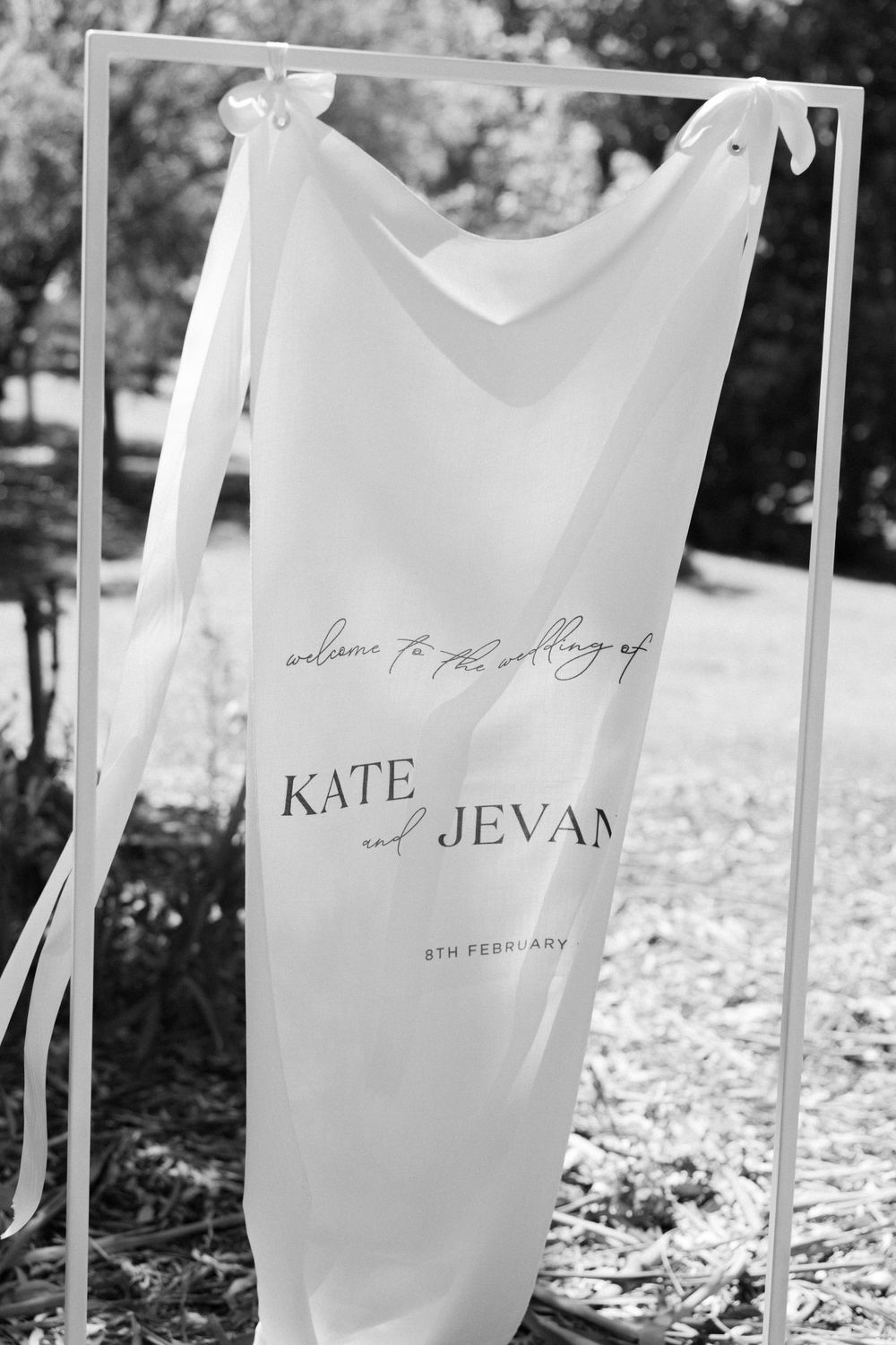 A black and white photo of a translucent wedding sign displaying 'Kate and Jevan' hanging from a white frame outdoors.