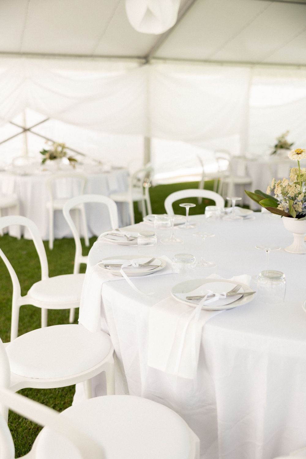 White round tables set for dining under an elegant tent with crisp linens and white chairs on grass flooring.