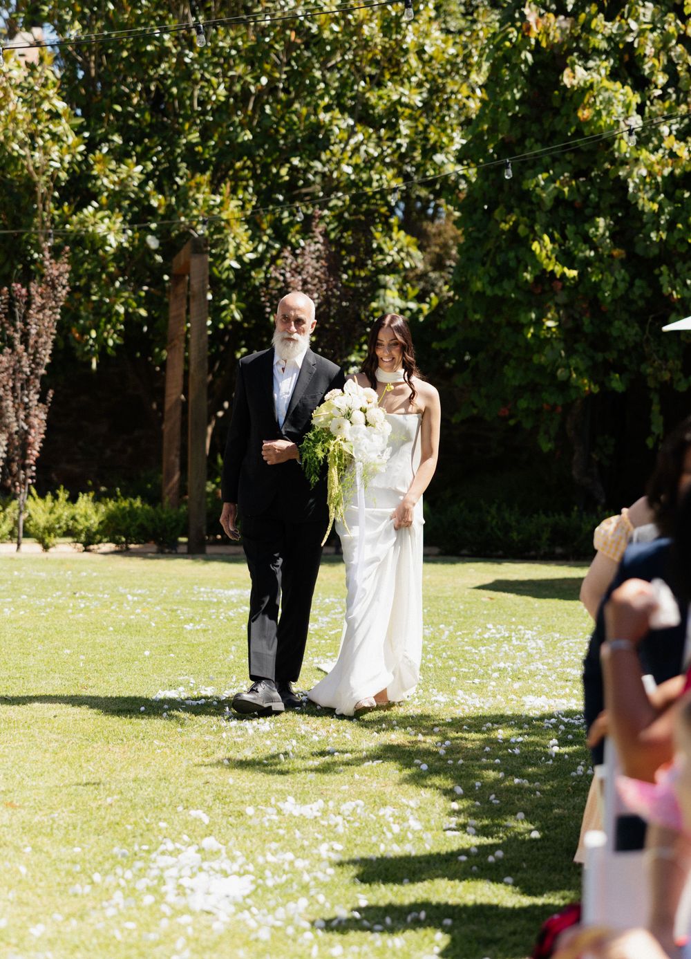 A wedding ceremony takes place on a sunlit lawn with green trees and flower petals scattered on the grass.