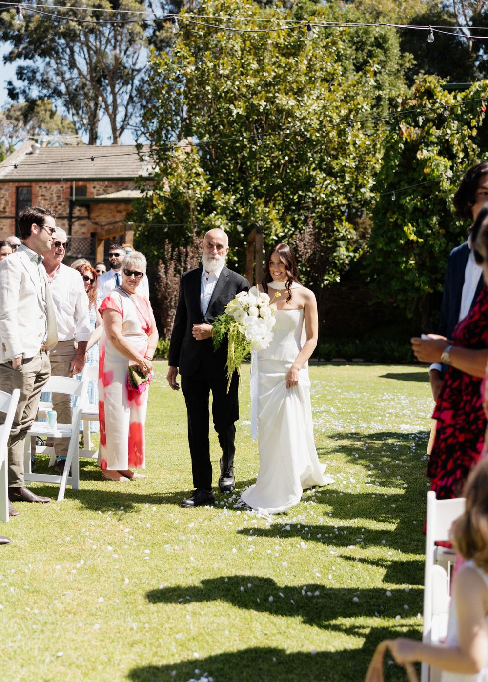 Outdoor wedding ceremony attendees gather on a sunny lawn with trees and a brick building visible in the background.