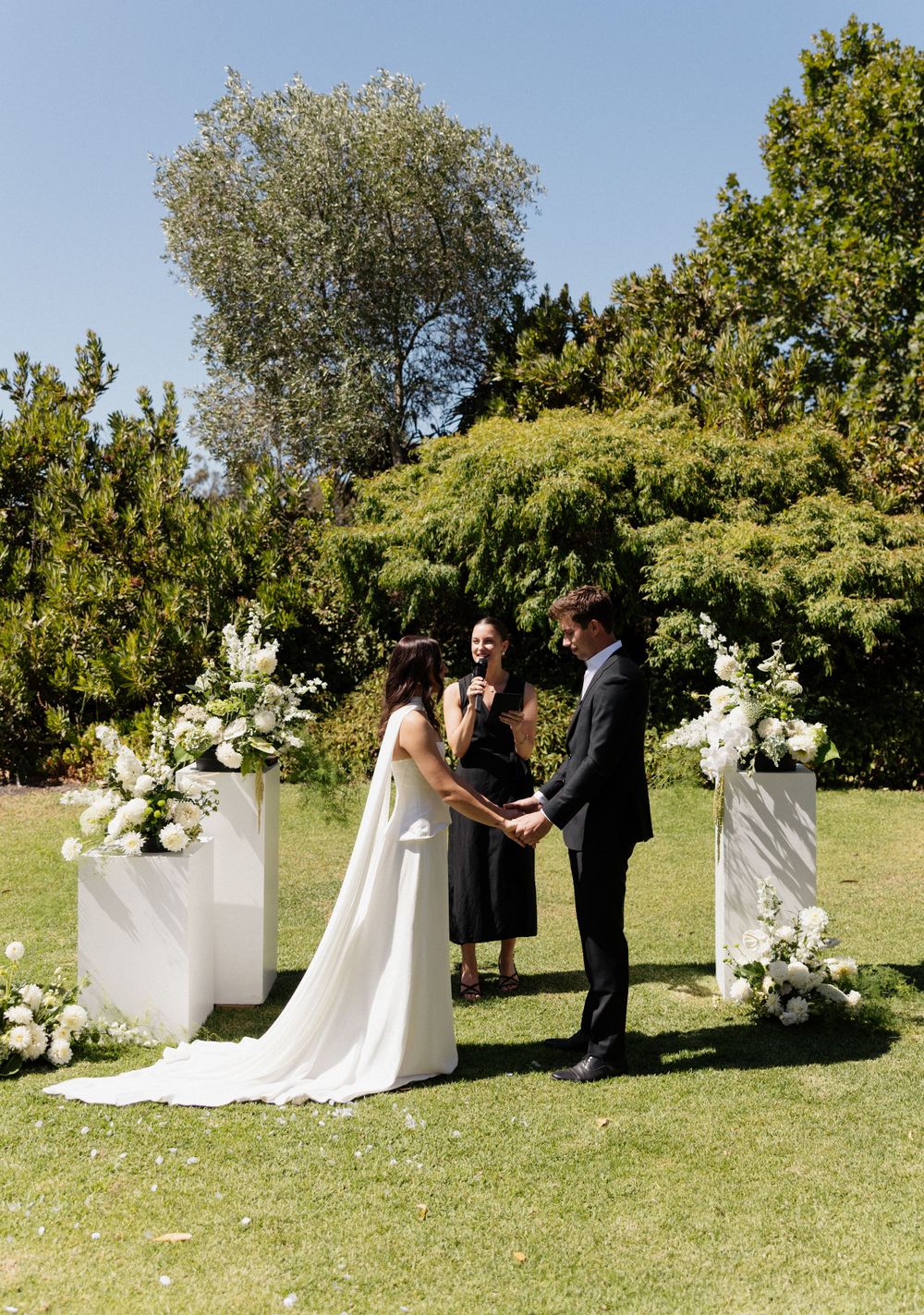 Garden wedding ceremony with white floral arrangements on pillars and lush trees creating a natural backdrop.