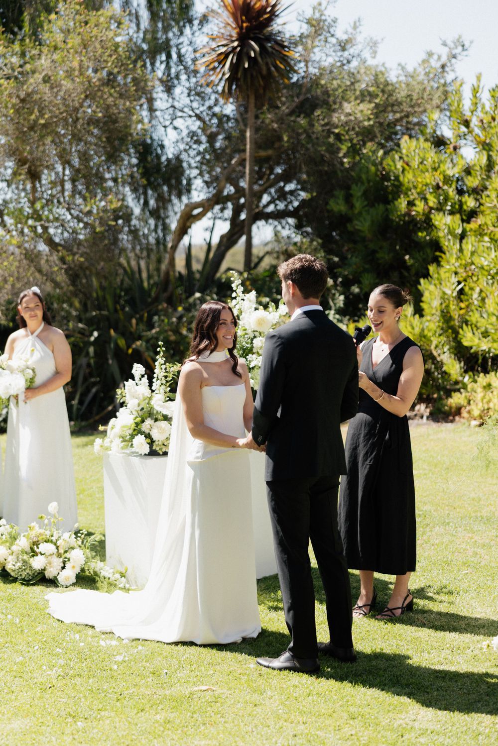 Wedding ceremony taking place outdoors on a sunny day surrounded by lush greenery and white floral arrangements.