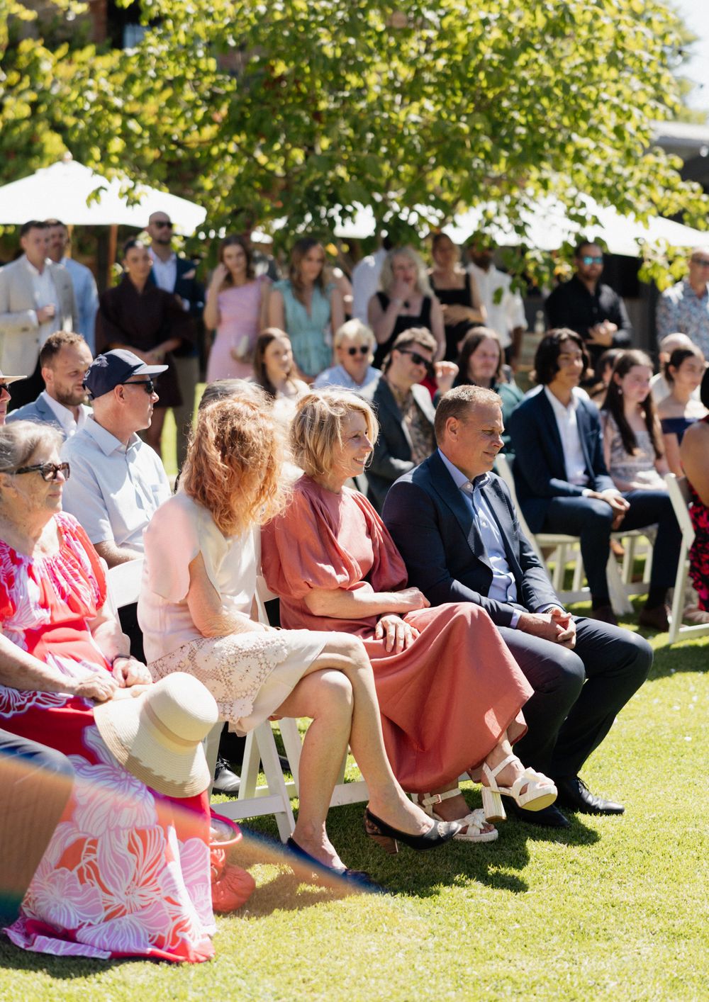 Wedding guests seated on white chairs on a sunny lawn during an outdoor ceremony with trees in the background.