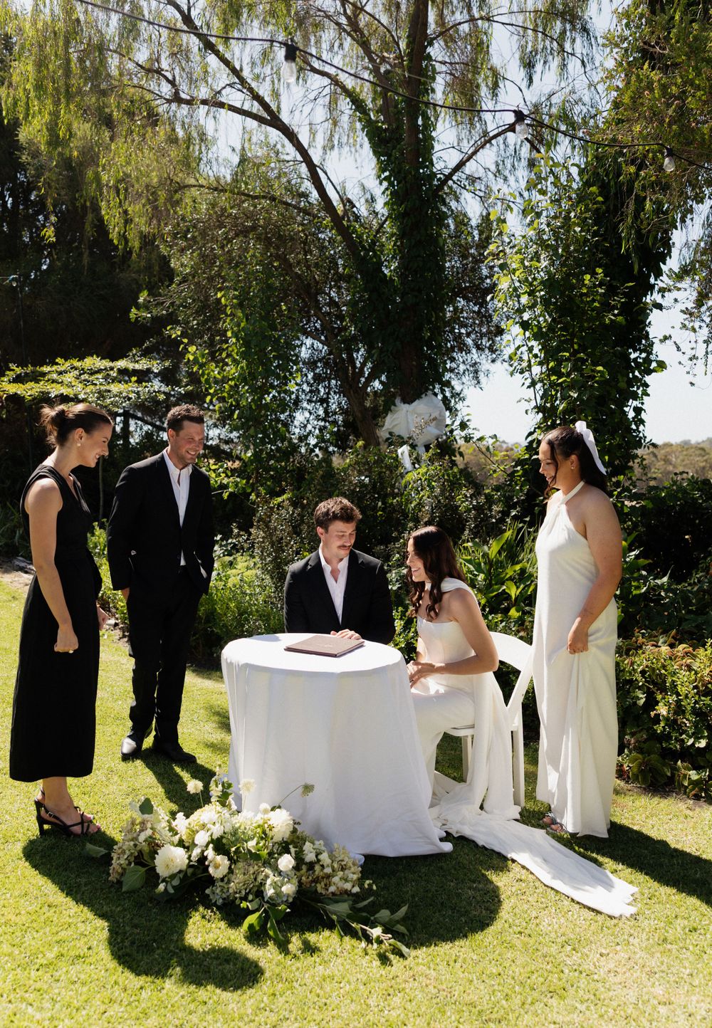 An outdoor wedding ceremony takes place in a sunny garden with white floral arrangements and a small signing table.