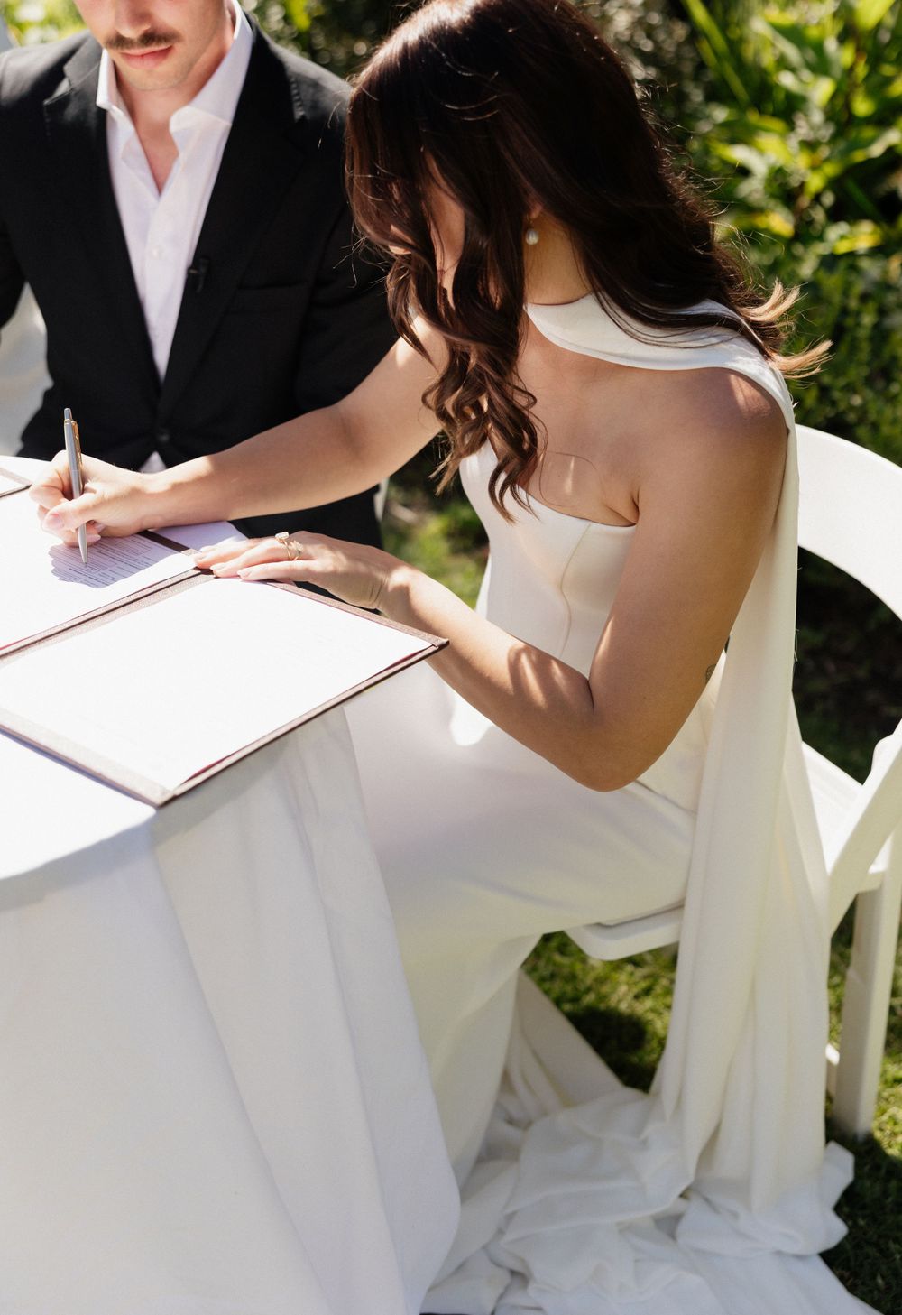 Couple signs marriage license at outdoor wedding ceremony with bride wearing elegant white strapless gown.