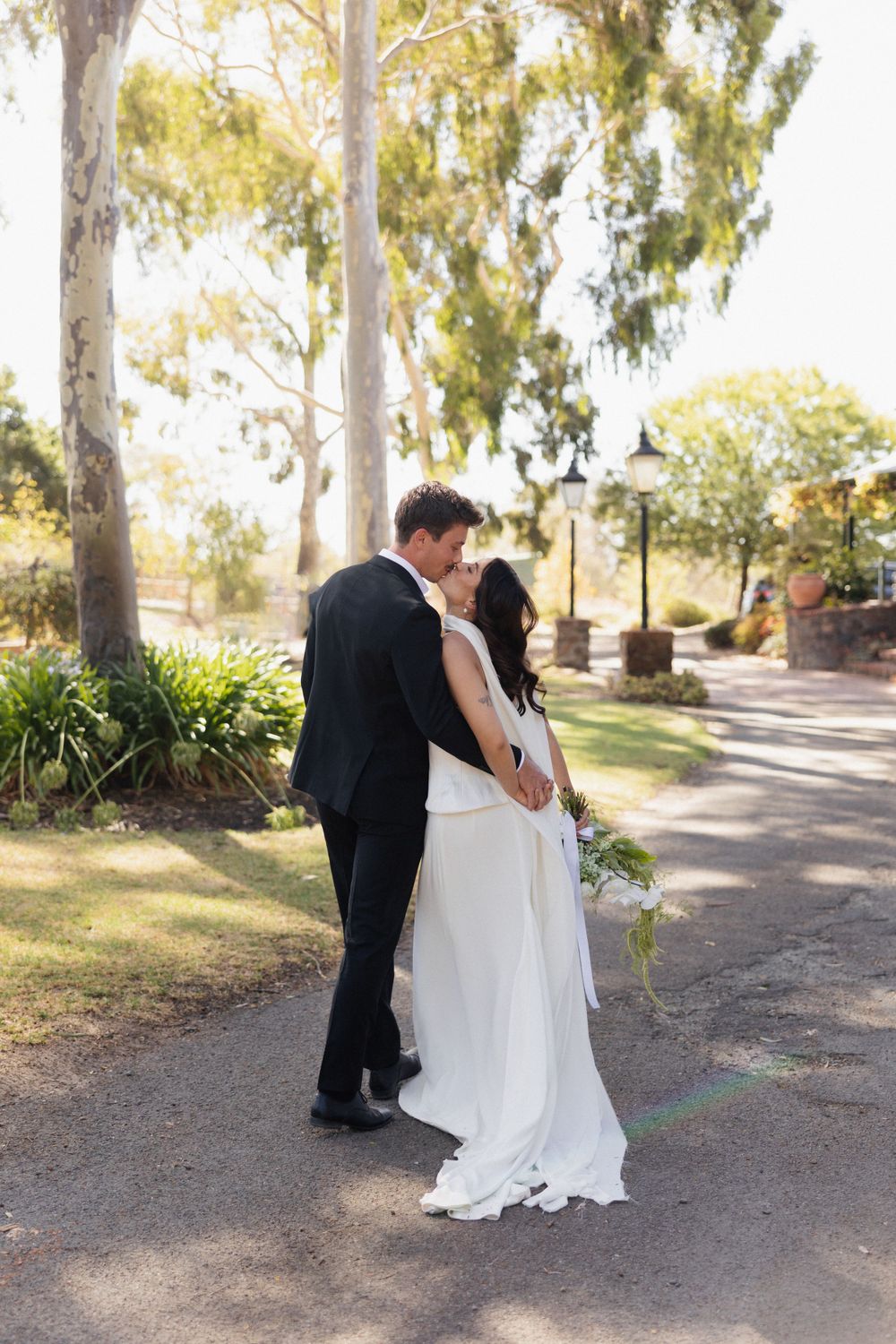 A couple embraces on a sunlit garden path surrounded by tall eucalyptus trees.