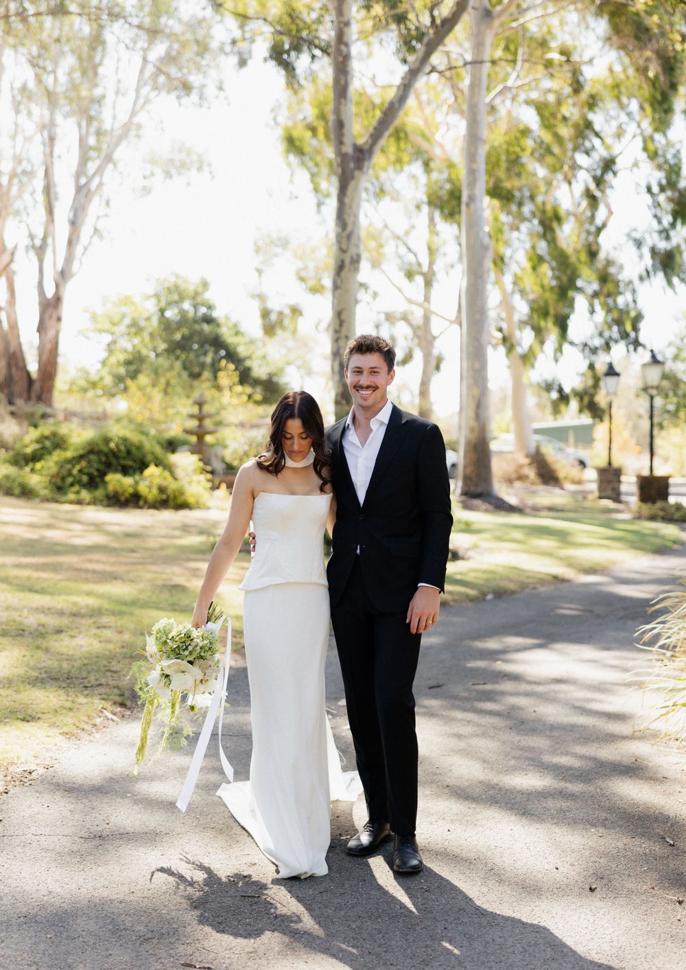 A bride and groom stand together on a sunlit path surrounded by tall eucalyptus trees in an outdoor garden setting.
