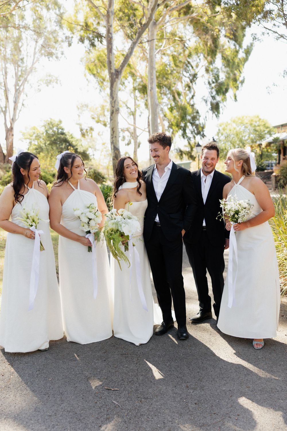 Wedding party in elegant white dresses and black suits pose together outdoors among eucalyptus trees on sunny day.