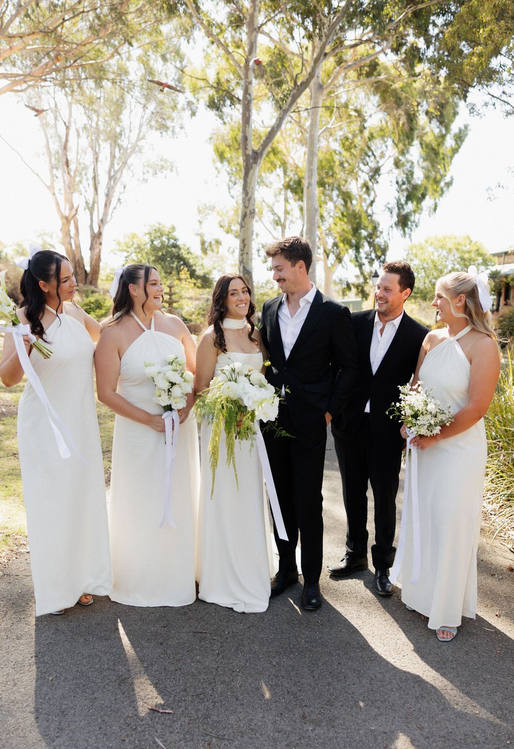 A wedding party wearing white dresses and black suits poses together on a sunlit path surrounded by trees.